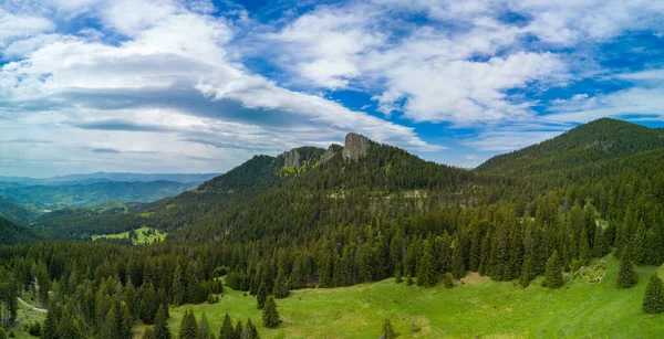 High rocky peak with mountain vegetation and dense spruce forests against blue daytime cloudy sky in valley of Rhodope Mountains. Panorama, top view