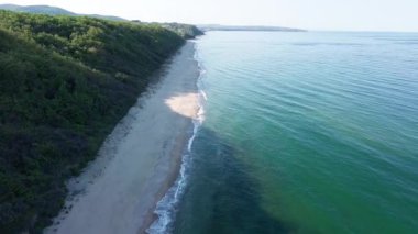 A wild empty sandy beach between a rocky sharp large ledge, covered with green fresh new vegetation, and a deep calm cool Black Sea under a clear blue sky. UHD 4K video realtime