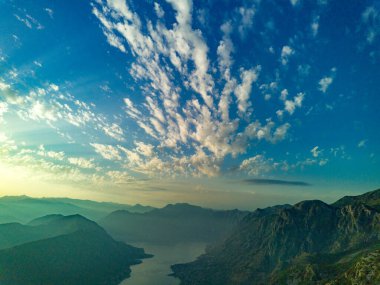 Bright dazzling golden summer sun in the blue cloudy evening sky illuminates all the silhouettes of the peaks of the Balkan Montenegrin mountains and the coast of the famous touristic Kotor Bay