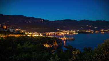 The large resort seaside town of Budva with hotels and fashionable beaches and boats with ships sail from coast to coast along the bay in light of bright night electric lights. UHD 4K video timelaps