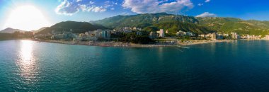 Panorama of sandy beach with sun umbrellas, plastic sunbeds and people relaxing in swimsuits in the historic tourist resort town of Becici near Adriatic Sea against the backdrop of evening sunny sky