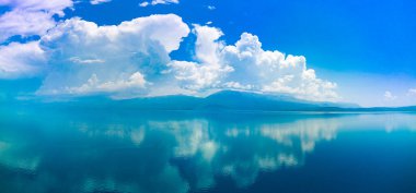Panorama of majestic huge white thick clouds float across the clear blue daytime sky above the horizon with the shallow azure calm Adriatic Sea in the light of the bright summer sun