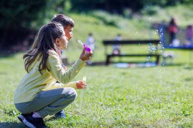 An older sister with long hair in a yellow tracksuit plays with her younger brother in a blue suit and teaches him how to blow soap bubbles on a green lawn in the backyard in spring