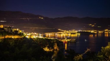 The large resort seaside town of Budva with hotels and fashionable beaches and boats with ships sail from coast to coast along the bay in light of bright night electric lights. UHD 8K video timelaps