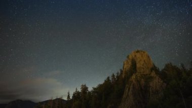 Rocky steep peak of a high mountain with dense dark spruce forests on the slopes against the backdrop of a magical starry clear night sky. UHD 8K video timelaps