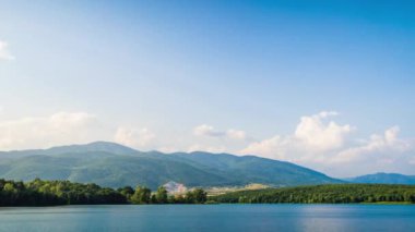 Small blue reflecting lake and clear cool water with stone narrow wild shore in coniferous spruce forest with tall fir trees, against pale blue daytime cloudy sky. UHD 8K video timelaps