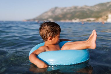 The baby sits in a blue inflatable small circle and looks for his mother, swimming in the blue warm summer Adriatic Sea in the evening sunlight