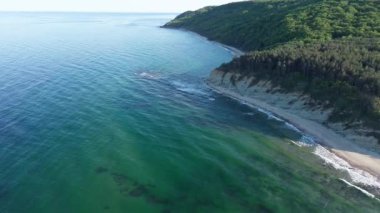 A wild empty sandy beach between a rocky sharp large ledge, covered with green fresh new vegetation, and a deep calm cool Black Sea under a clear blue sky. UHD 4K video realtime