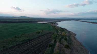 Calm bluish water surface of Black Sea washes wild coast and green farmer fields in valley of Bulgaria, against background of cloudy color vast sky with bright sunset sun. UHD 4K video realtime