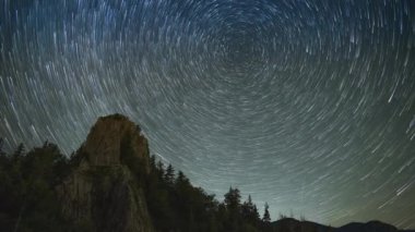 Rocky steep peak of a high mountain with dense dark spruce forests on the slopes against the backdrop of a magical starry clear night sky. UHD 4K video timelaps