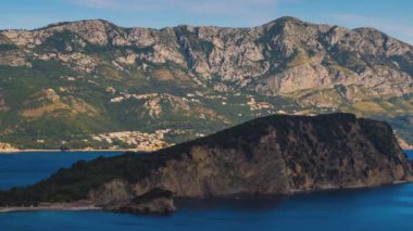 A small historical island of St. Nichola in the calm warm Adriatic Sea near the shores of the bustling tourist resort town of Budva in the rays of the evening sunset summer sun. UHD 4K video timelaps