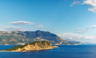 Historical ancient tourist island of St. Nicholas with vegetation on rocky shores in the azure Adriatic Sea against the backdrop of coastal resort towns, the Montenegrin mountain range and clear sky