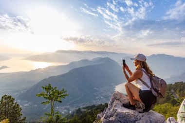 A happy contented young girl with a backpack takes a souvenir photo of the Montenegrin coastal cities, the Adriatic Sea and the bright sunset rays from the top of a high mountain