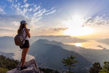 A happy contented young girl with a backpack takes a souvenir photo of the Montenegrin coastal cities, the Adriatic Sea and the bright sunset rays from the top of a high mountain