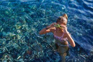 A small thin wet girl in a bright childrens swimsuit with blue swimming goggles on her head is going to dive onto the rocky colorful bottom of the transparent warm summer Adriatic Sea