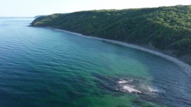 A wild empty sandy beach between a rocky sharp large ledge, covered with green fresh new vegetation, and a deep calm cool Black Sea under a clear blue sky. UHD 4K video realtime