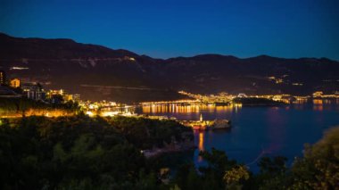 The large resort seaside town of Budva with hotels and fashionable beaches and boats with ships sail from coast to coast along the bay in light of bright night electric lights. UHD 8K video timelaps