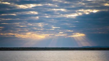Bright sunset rays of the sun shine through thick evening white clouds over the blue calm Black Sea. UHD 4k video timelaps