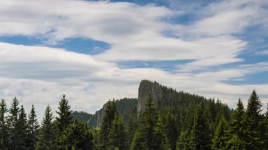 Rocky sharp high peak of one of the Rhodope Mountains with a sheer cliff and a steep slope with a dark evergreen spruce forest against the background of a daytime blue cloudy sky. UHD 4K video