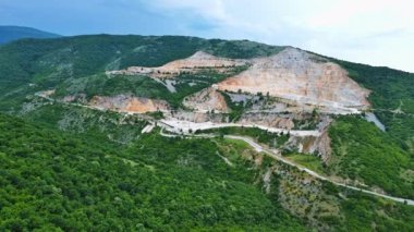 Natural stepped quarry rich in minerals is located near winding intermountain road, against backdrop of Rhodope Mountains and hills with evergreen spruce forests and vegetation. UHD 4K video realtime