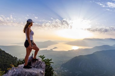 Happy satisfied young girl with a backpack looks at the coastal sea Montenegrin cities, the Adriatic Sea and the bright sunset rays from the top of a high mountain