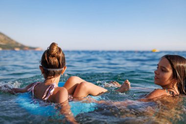 An older sister with dark hair rides her younger sister with blue swimming goggles, who is sitting in a small bright inflatable ring in the warm waters of the Adriatic Sea in the evening sunlight