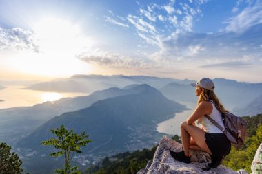 Happy satisfied young girl with a backpack looks at the coastal sea Montenegrin cities, the Adriatic Sea and the bright sunset rays from the top of a high mountain