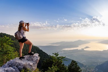 A happy contented young girl with a backpack takes a souvenir photo of the Montenegrin coastal cities, the Adriatic Sea and the bright sunset rays from the top of a high mountain