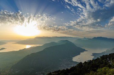 Bright dazzling golden summer sun in the blue cloudy evening sky illuminates all the silhouettes of the peaks of the Balkan Montenegrin mountains and the coast of the famous touristic Kotor Bay