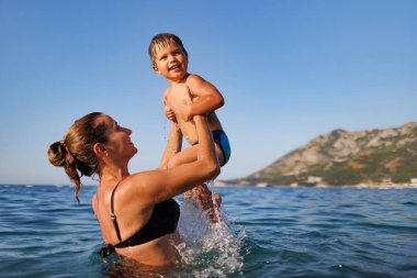 A happy mother in a bathing suit holds her little son in her arms and throws him over her head in the warm blue Adriatic Sea in the evening sunlight