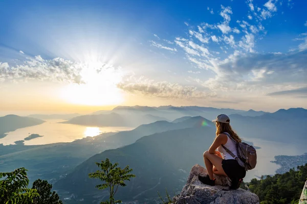 Happy satisfied young girl with a backpack looks at the coastal sea Montenegrin cities, the Adriatic Sea and the bright sunset rays from the top of a high mountain