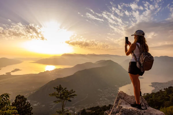 A happy contented young girl with a backpack takes a souvenir photo of the Montenegrin coastal cities, the Adriatic Sea and the bright sunset rays from the top of a high mountain