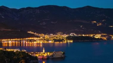 The large resort seaside town of Budva with hotels and fashionable beaches and boats with ships sail from coast to coast along the bay in light of bright night electric lights. UHD 4K video timelaps