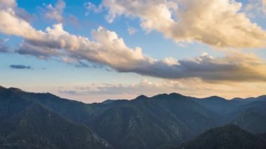Unusual white fluffy clouds float across the blue evening sky with sunlight over the mountain valley of the Rhodope Mountains with dense dark spruce forests on the slopes. UHD 4K video timelaps