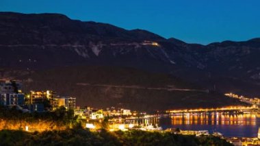 The large resort seaside town of Budva with hotels and fashionable beaches and boats with ships sail from coast to coast along the bay in light of bright night electric lights. UHD 4K video timelaps