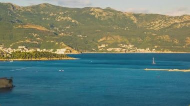 Top view of resort seaside town of Budva with various holiday hotels and fashionable beaches and boats with ships sail from coast to coast along bay in the light of setting sun. UHD 4K video timelaps