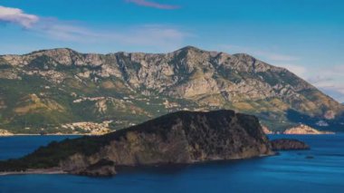 A small historical island of St. Nichola in the calm warm Adriatic Sea near the shores of the bustling tourist resort town of Budva in the rays of the evening sunset summer sun. UHD 4K video timelaps