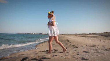 Ukraynalı beyaz gömlekli, çiçekli Ukrayna çelengi giyen, çok renkli, parlak kurdeleli, günbatımında Karadeniz kıyısında dans eden küçük bir kız. UHD Video yavaş çekim 4K