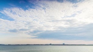 A light wind shakes dry reeds and makes ripples on a small bay of a calm river with a reflection of a cloudy blue sky. UHD 4k video timelaps