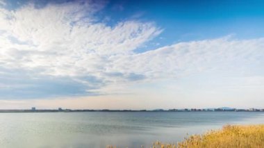 A light wind shakes dry reeds and makes ripples on a small bay of a calm river with a reflection of a cloudy blue sky. UHD 4k video timelaps