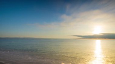 Calm blue boundless Black Sea with cool water reflecting light and sandy wild empty beach nearby, against cloudy colored sky with white clouds and bright dawn sun. UHD 4K video timelaps