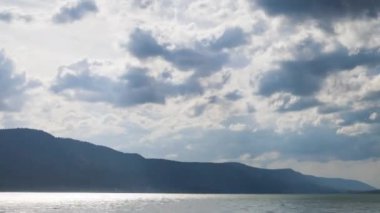 Small blue reflecting lake and clear cool water with stone narrow wild shore in coniferous spruce forest with tall fir trees, against pale blue daytime cloudy sky. UHD 4K video timelaps