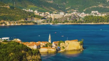 Top view of resort seaside town of Budva with various holiday hotels and fashionable beaches and boats with ships sail from coast to coast along bay in the light of setting sun. UHD 4K video timelaps