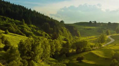 A winding asphalt intermountain road passes high green hills covered with spruce forests and fresh spring wide meadows against a cloudy gray boundless sky. UHD 4K video timelaps
