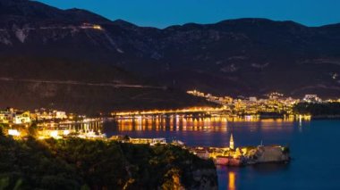 The large resort seaside town of Budva with hotels and fashionable beaches and boats with ships sail from coast to coast along the bay in light of bright night electric lights. UHD 4K video timelaps