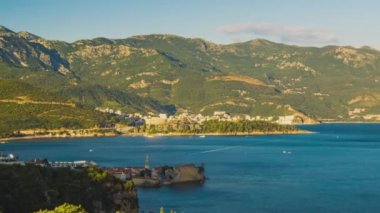 Top view of resort seaside town of Budva with various holiday hotels and fashionable beaches and boats with ships sail from coast to coast along bay in the light of setting sun. UHD 4K video timelaps