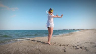 Ukraynalı beyaz gömlekli, çiçekli Ukrayna çelengi giyen, çok renkli, parlak kurdeleli, günbatımında Karadeniz kıyısında dans eden küçük bir kız. UHD Video yavaş çekim 4K