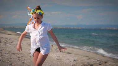 Ukraynalı beyaz gömlekli, çiçekli Ukrayna çelengi giyen, çok renkli, parlak kurdeleli, günbatımında Karadeniz kıyısında dans eden küçük bir kız. UHD Video yavaş çekim 4K