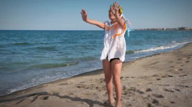 Ukraynalı beyaz gömlekli, çiçekli Ukrayna çelengi giyen, çok renkli, parlak kurdeleli, günbatımında Karadeniz kıyısında dans eden küçük bir kız. UHD Video yavaş çekim 4K