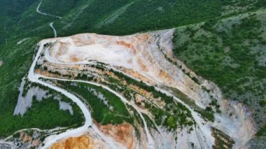 Natural stepped quarry rich in minerals is located near winding intermountain road, against backdrop of Rhodope Mountains and hills with evergreen spruce forests and vegetation. UHD 4K video realtime